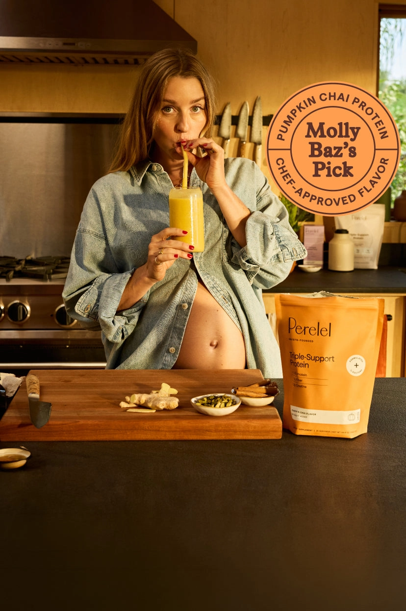 A woman in a denim jacket drinking a Pumpkin Chai protein drink in a kitchen